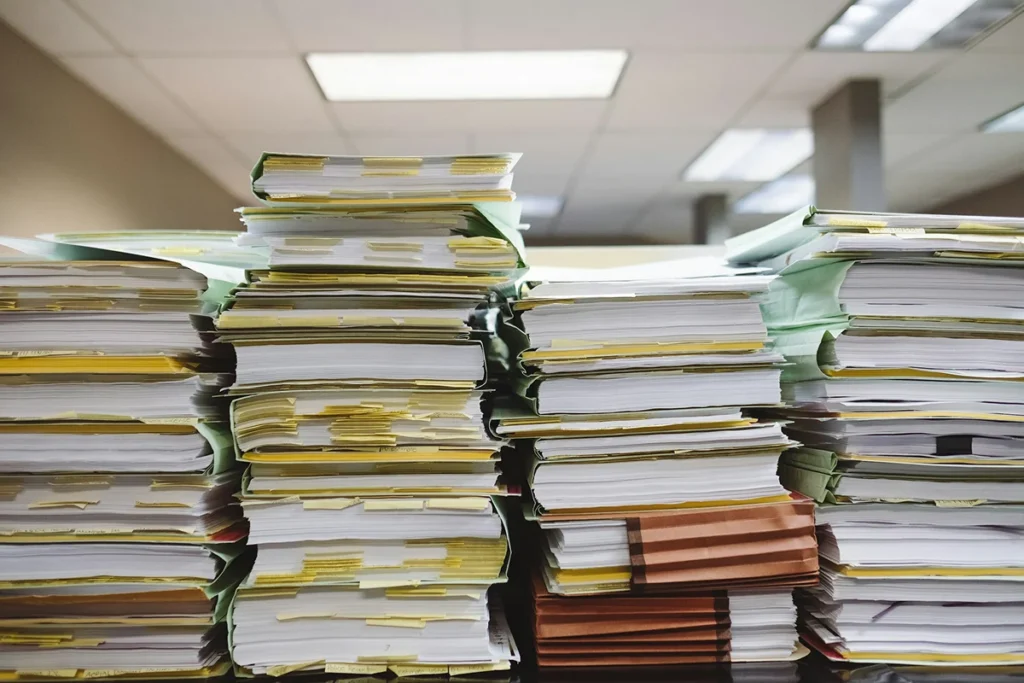 A large stack of paper files and folders is shown, representing the extensive documentation and potential for inefficiency associated with government and bureaucratic processes.
