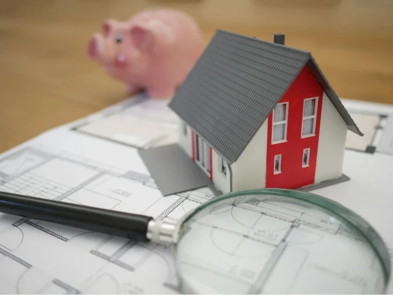 A small model of a house and a piggy bank sit on top of architectural blueprints, representing the investment and planning involved in real estate.